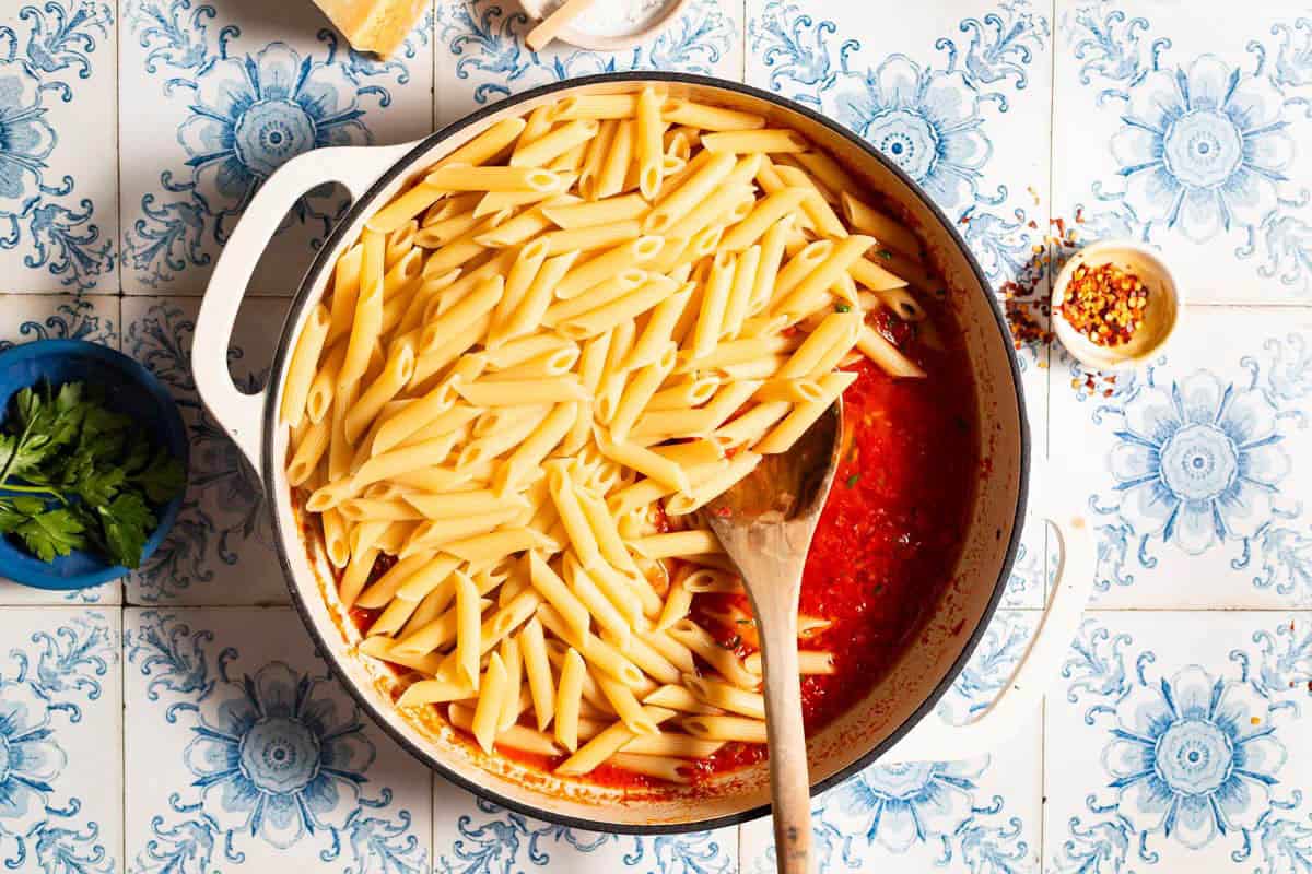 Penne pasta being added to arrabbiata sauce in a skillet with a wooden spoon. Next to this are small bowls of red pepper flakes and parsley.