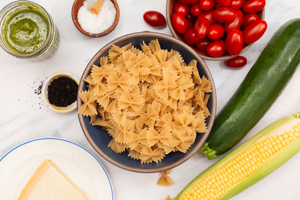 Ingredients for summer pasta salad including bowtie pasta, salt, zucchini, corn, urfa biber, cherry tomatoes, parmesan, and basil vinaigrette.