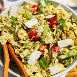 A close up of summer pasta salad in a serving bowl with wooden serving utensils. Next to this is a bowl of urfa biber, a plate of parmesan cheese and a cloth napkin.