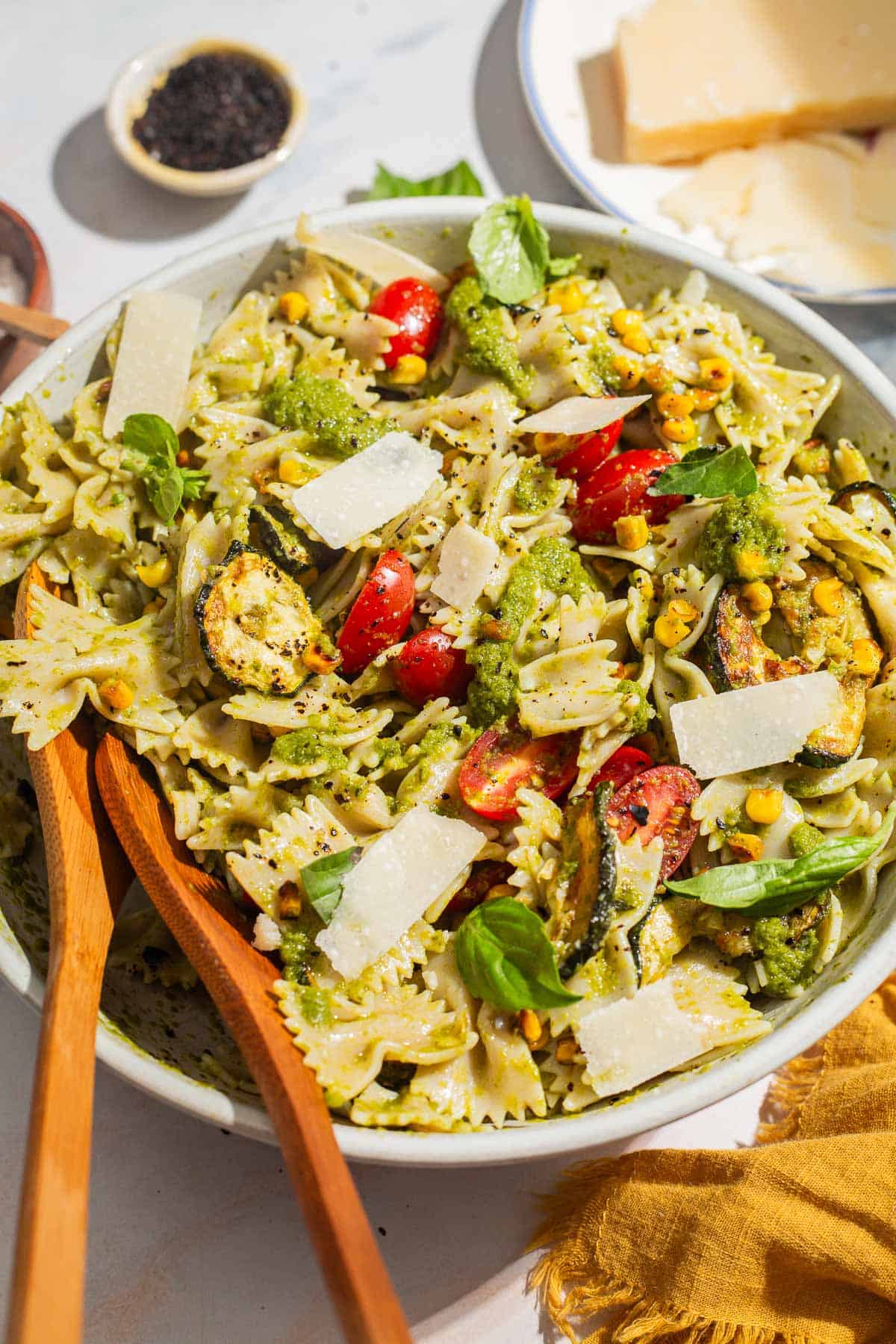 A close up of summer pasta salad in a serving bowl with wooden serving utensils. Next to this is a bowl of urfa biber, a plate of parmesan cheese and a cloth napkin.