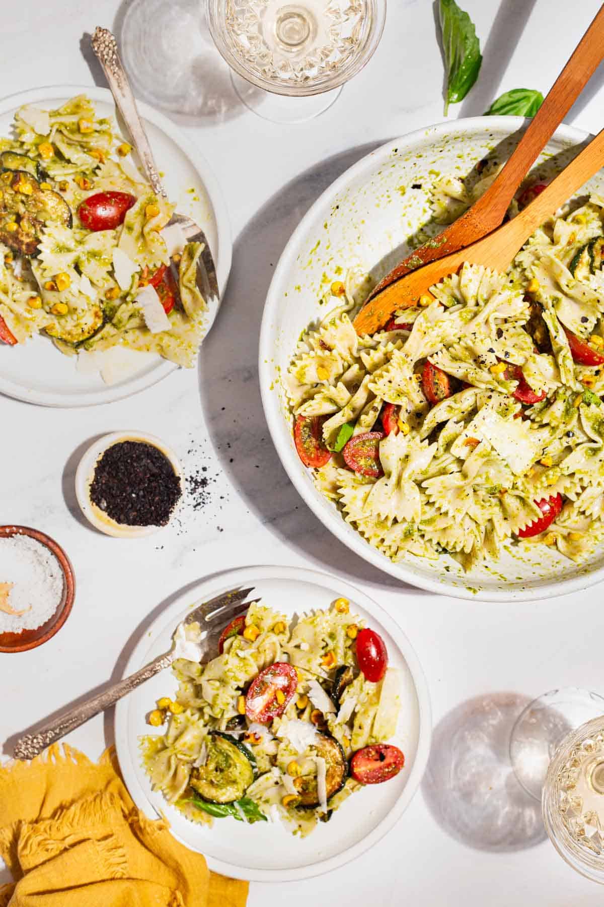 An overhead photo of 2 servings of summer pasta salad on plates with forks. Surrounding these are 2 glasses of white wine, a serving bowl of the pasta salad with wooden serving utensils, bowls of urfa biber and salt, and a cloth napkin.