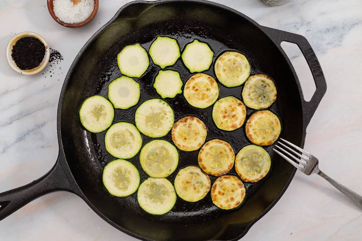 An overhead photo of slices of zucchini being sauteed in a cast iron skillet. Next to this is a fork and bowls of salt and urfa biber.