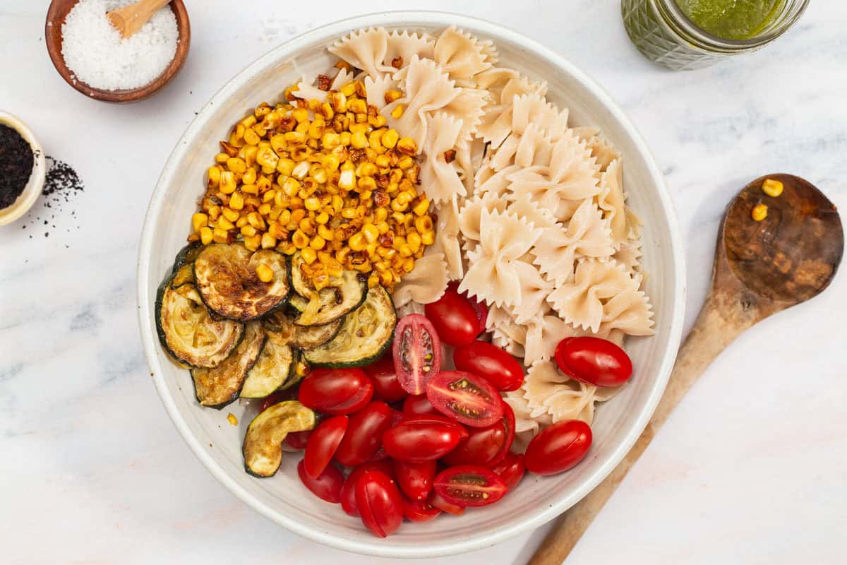 Cooked pasta, zucchini and corn along with halved cherry tomatoes in a bowl just before being mixed together. Next to his is a wooden spoon, bowls of urfa biber and salt, and a jar of basil vinaigrette.