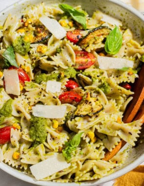 A close up of summer pasta salad in a serving bowl with wooden serving utensils. Next to this is a bowl of urfa biber, and a cloth napkin.