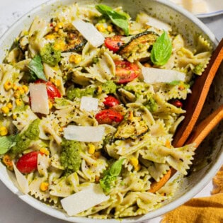 A close up of summer pasta salad in a serving bowl with wooden serving utensils. Next to this is a bowl of urfa biber, and a cloth napkin.