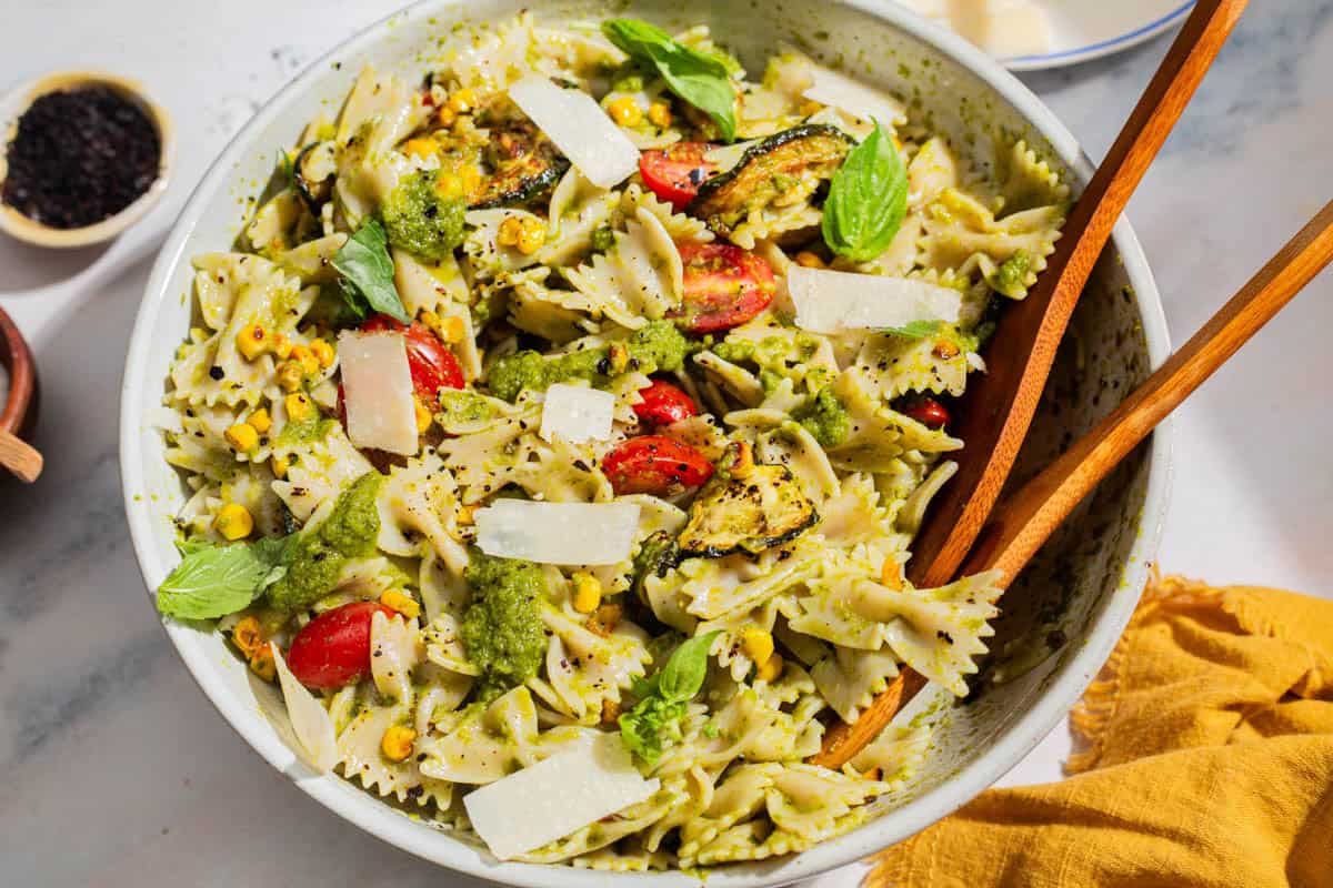 A close up of summer pasta salad in a serving bowl with wooden serving utensils. Next to this is a bowl of urfa biber, and a cloth napkin.