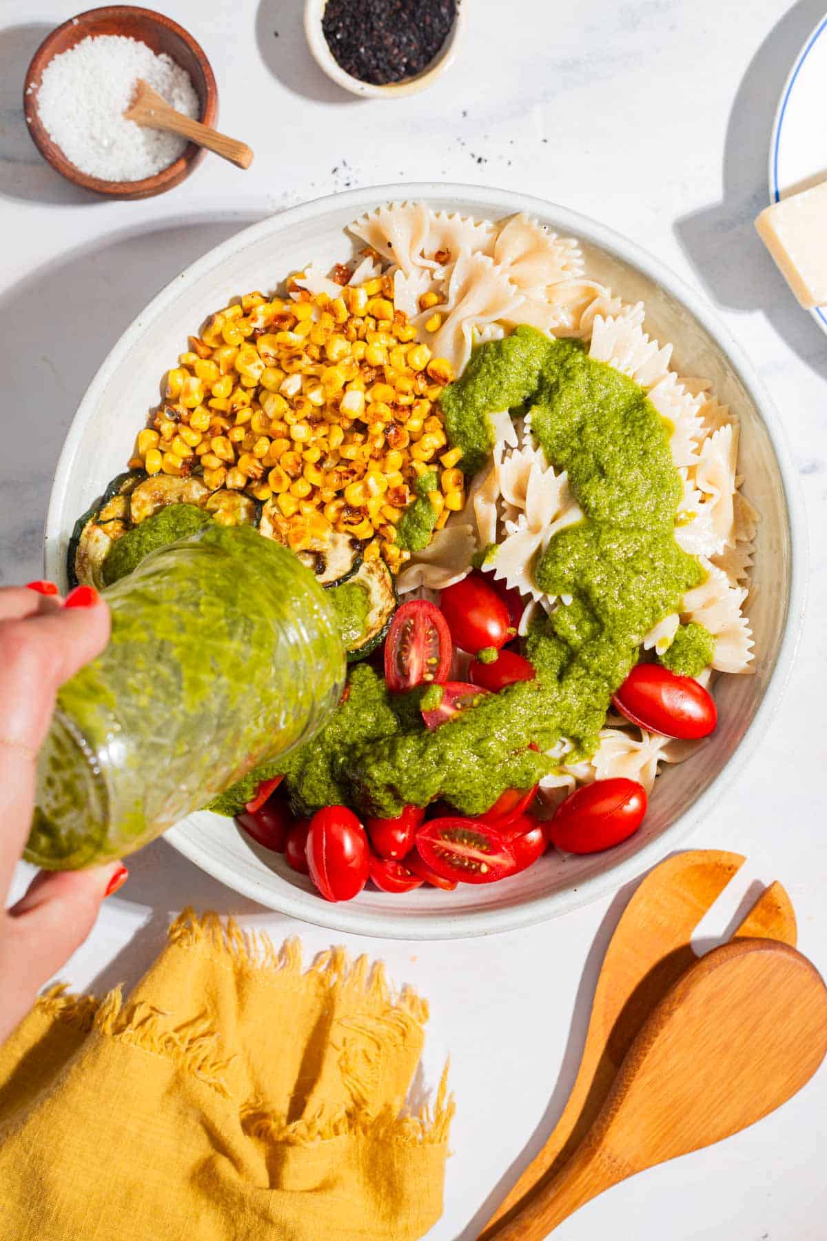 An overhead photo of the basil vinaigrette being poured onto the unmixed summer pasta salad in a bowl. Next to this is a cloth napkin, wooden serving utensils, and bowls of salt and urfa biber.