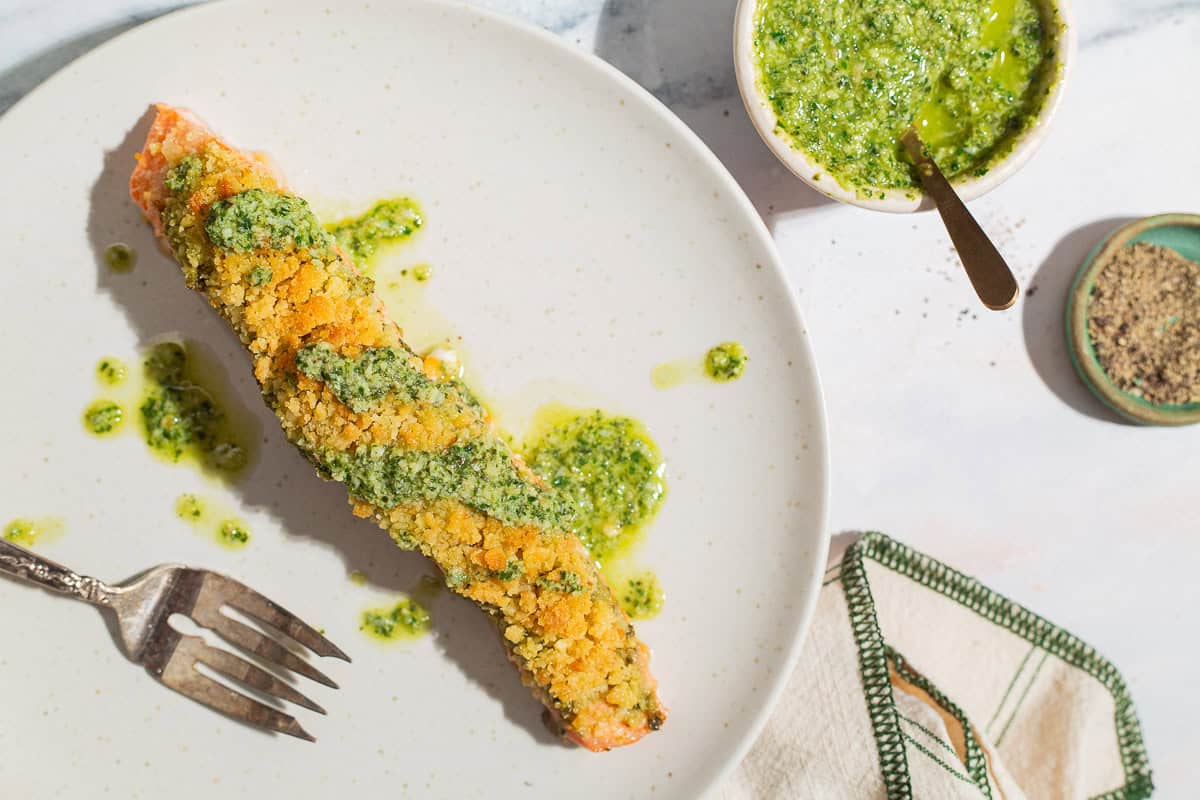 An overhead photo of a pesto salmon fillet on a plate with a fork. Next to this is a bowl of pesto with a fork, a bowl of black pepper and a cloth napkin.