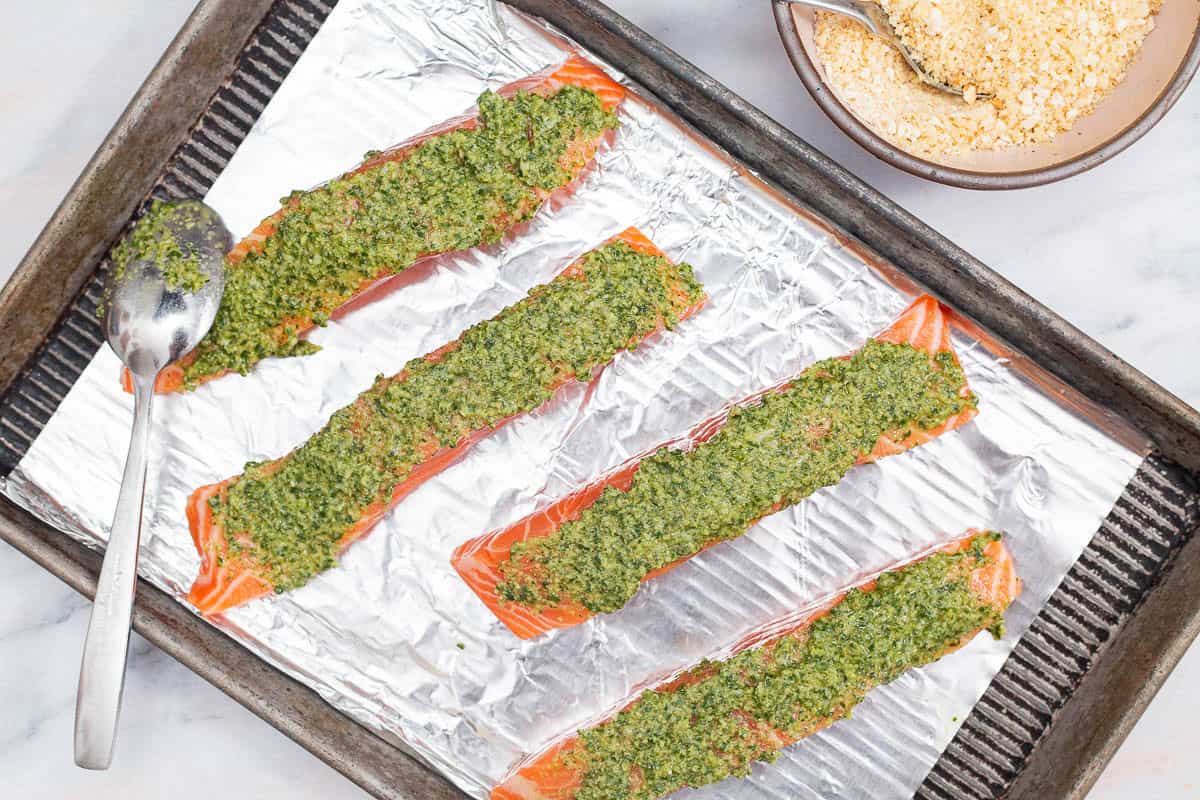 An overhead photo of 4 uncooked salmon fillets topped with pesto an panko on a baking sheet lined with aluminum foil and a spoon. Next to this is a bowl of breadcrumbs with a spoon.