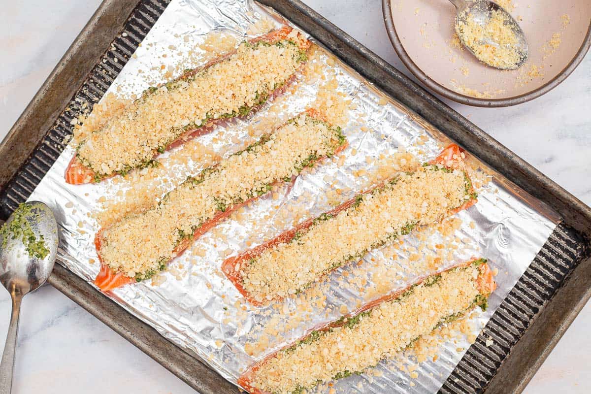 An overhead photo of 4 uncooked salmon fillets topped with pesto an panko bread crumbs on a baking sheet lined with aluminum foil and a spoon. Next to this is an empty bowl with a spoon.