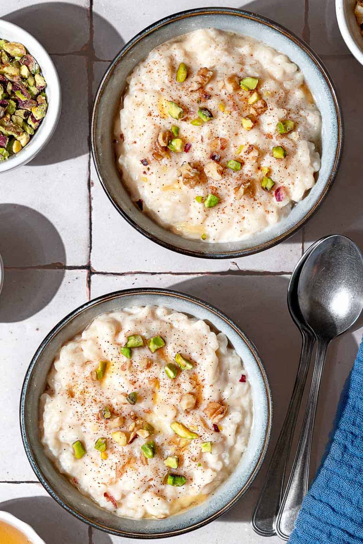 An overhead photo of 2 bowls of easy rice pudding garnished with pistachios, walnuts, cinnamon and a drizzle of honey. Next to this is a kitchen towel, 2 spoons and a bowl of crushed pistachios.