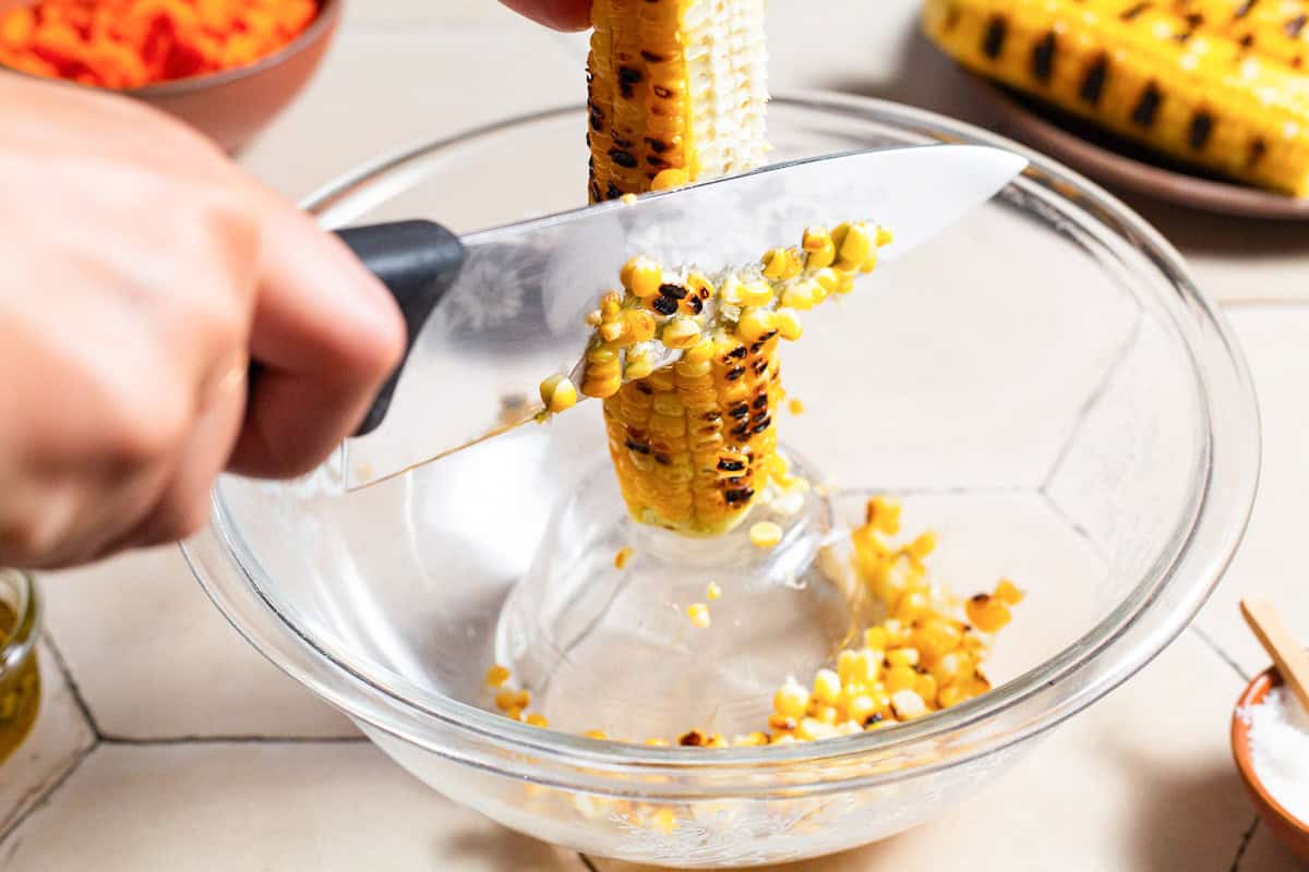 Grilled corn being cut from a cob in a bowl with a knife.
