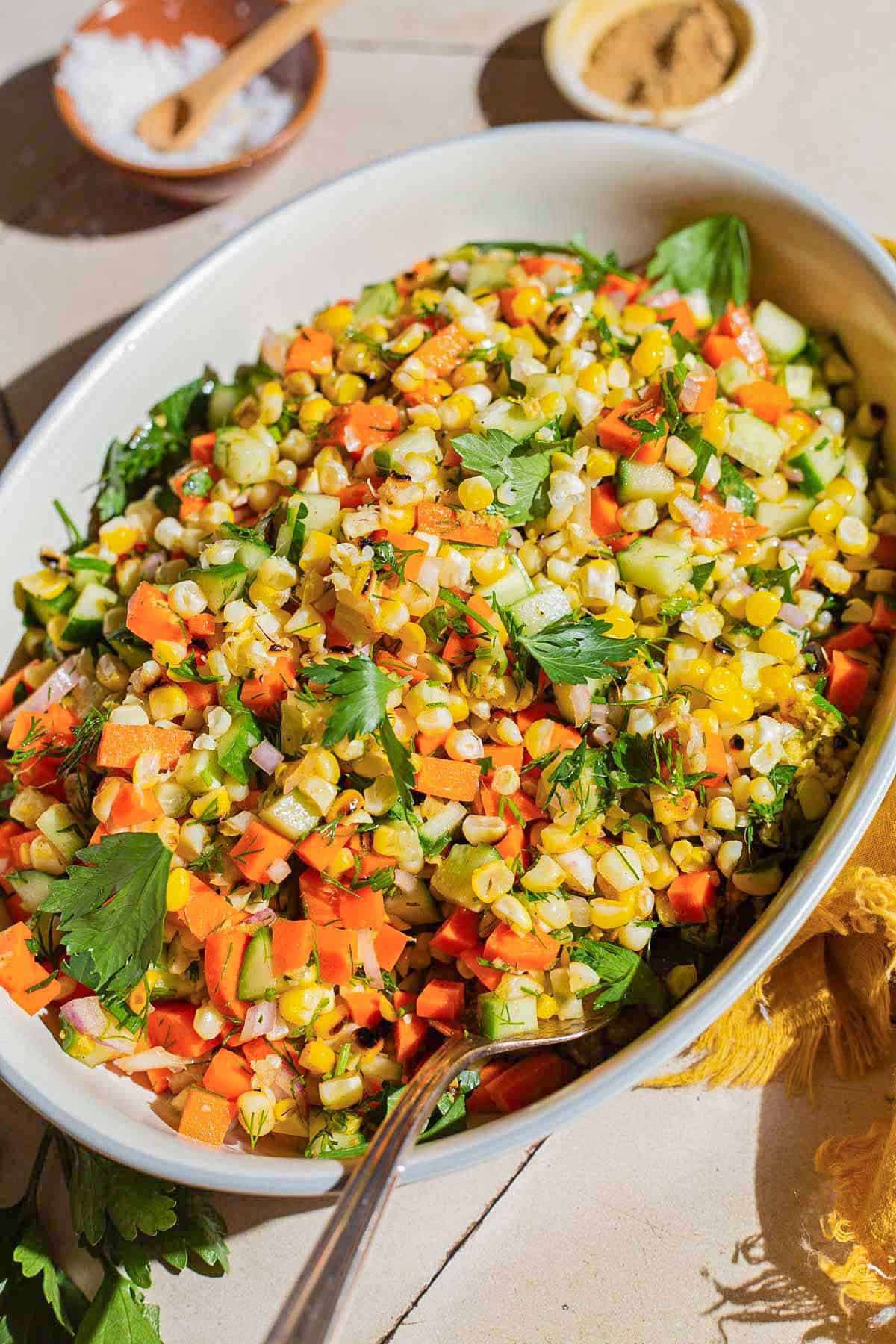 Grilled corn salad in a serving bowl with a spoon. Next to this are small bowls of salt and cumin.