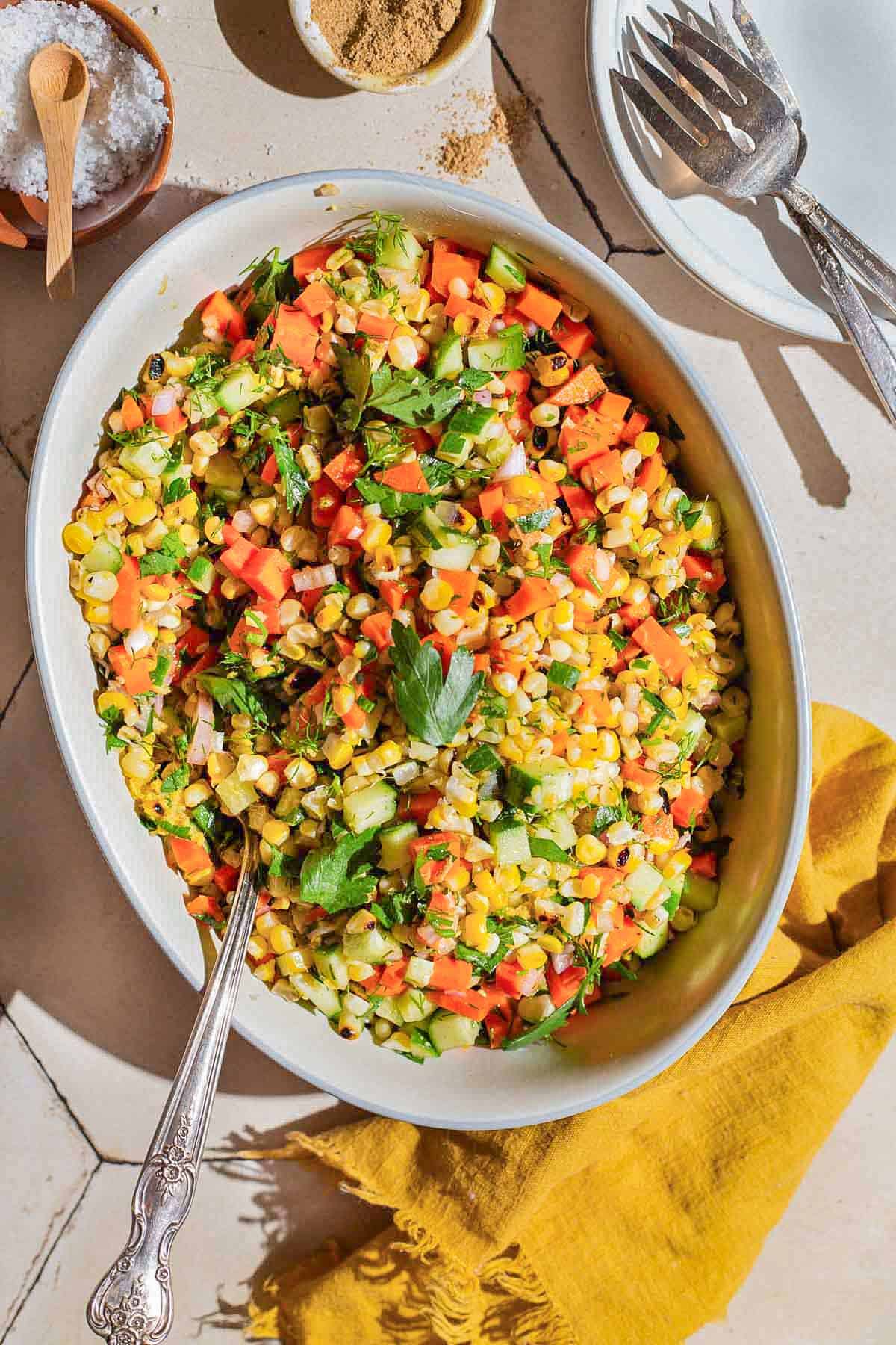 An overhead photo of grilled corn salad in a serving bowl with a spoon. Next to this is a cloth napkin, small bowls of salt and cumin, and a plate with 2 forks.