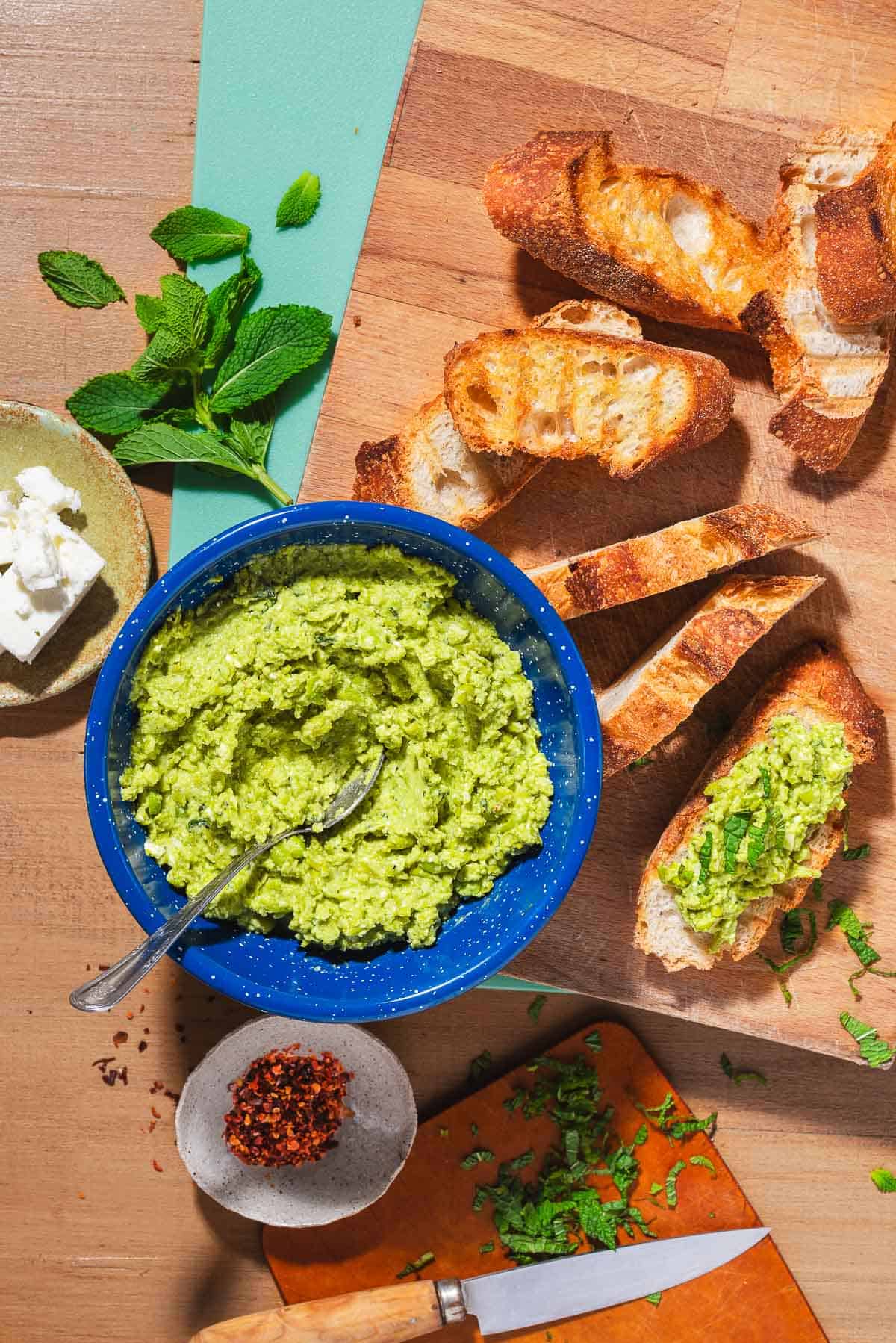 An overhead photo of a bowl of smashed peas with a spoon, several pieces of the toasted baguette, a sprig of fresh mints, bowls of feta and aleppo pepper and a knife.