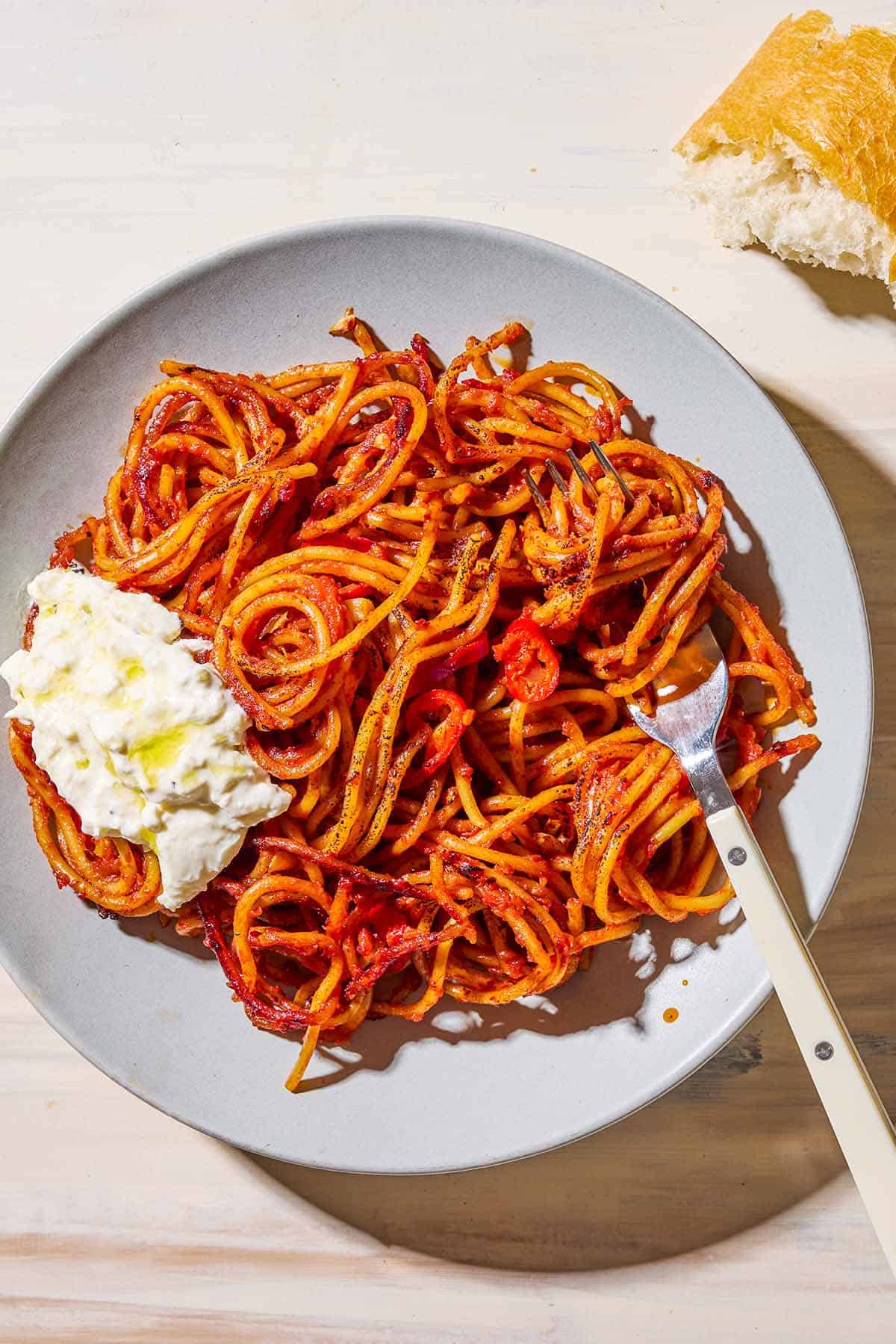 An overhead photo of a serving spaghetti all'assassina topped with burrata on a plate with a fork. Next to this is a slice of crusty bread.