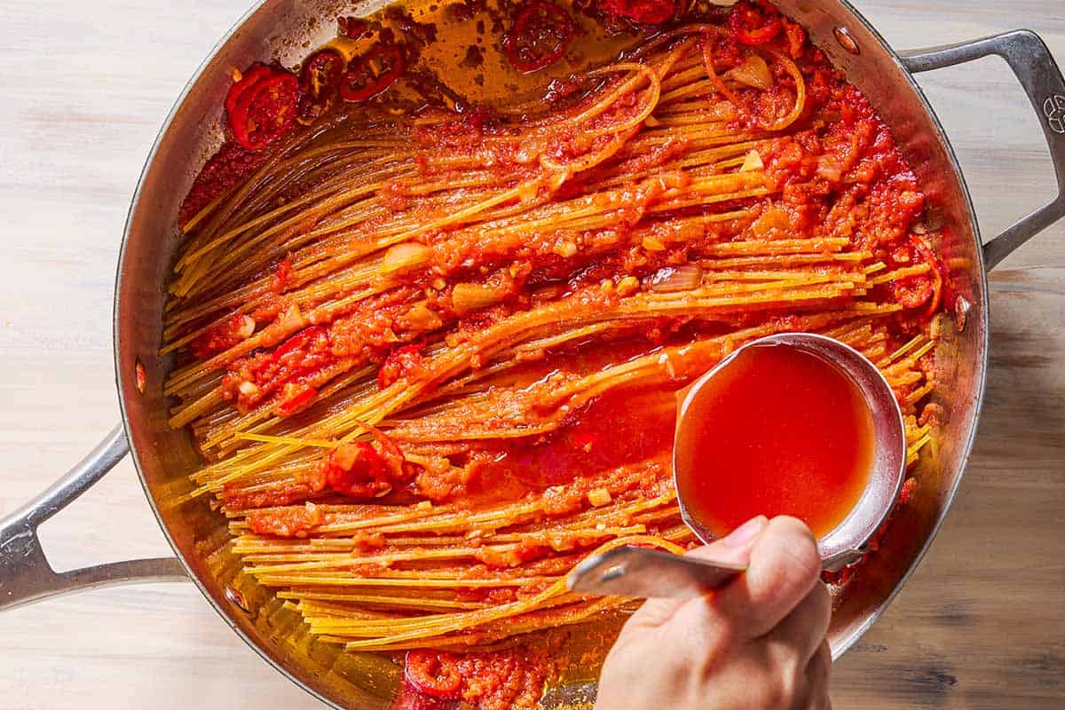 An overhead photo of the tomato sauce being added to the cooking spaghetti all'assassina in a skillet with a ladle.