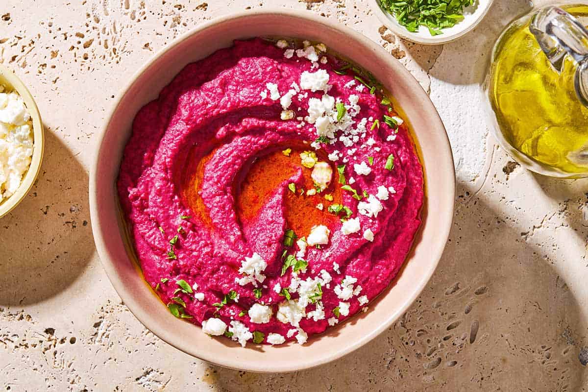 An overhead photo of beet hummus in a bowl topped with olive oil, feta cheese and parsley. Next to this is a bottle of olive oil and bowls of parsley and feta.