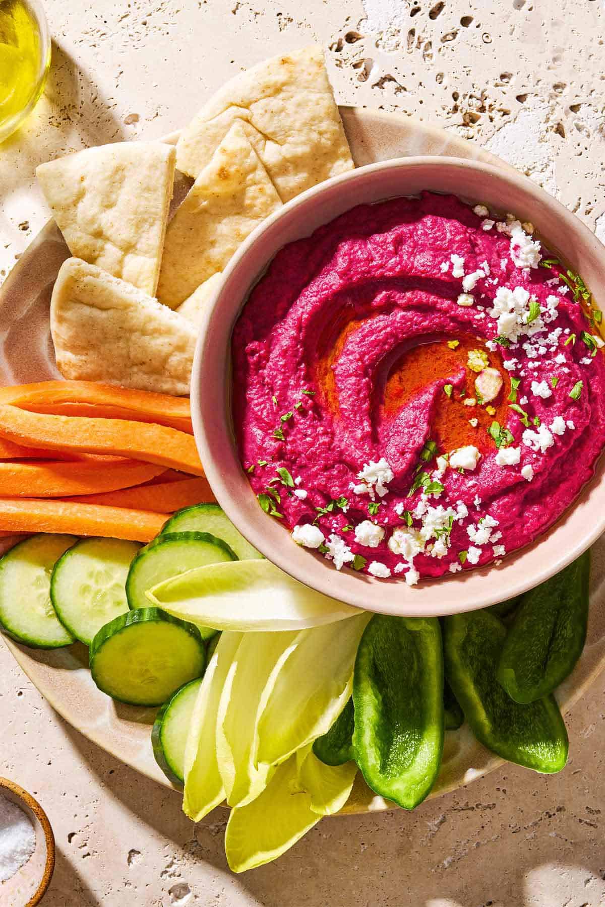 An overhead photo of beet hummus in a bowl topped with olive oil, feta cheese and parsley on a platter with pita wedges and various sliced vegetables.