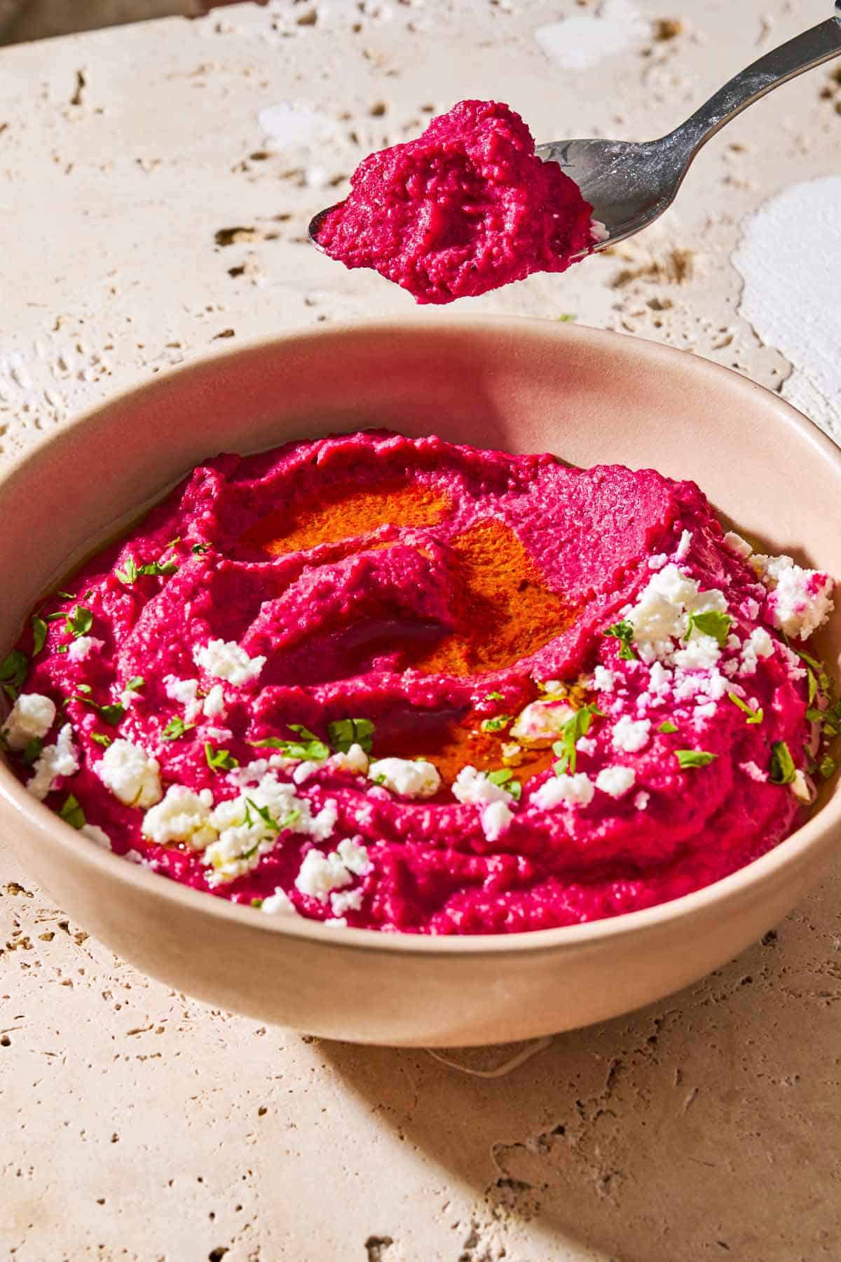 A spoonful of beet hummus being lifted from a bowl of beet hummus in a bowl topped with olive oil, feta cheese and parsley.