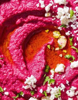A close up of beet hummus in a bowl topped with olive oil, feta cheese and parsley.