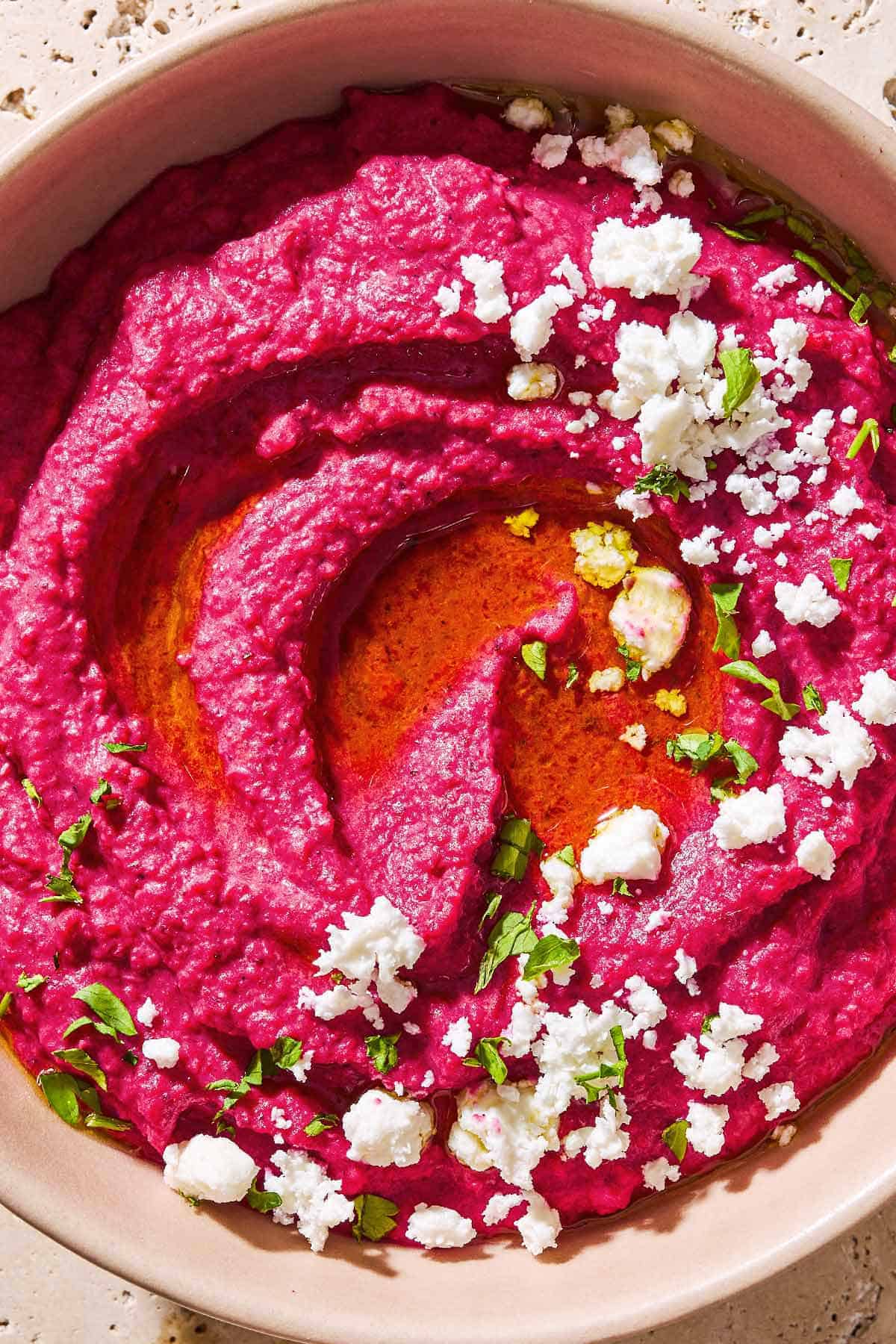 An overhead close up photo of beet hummus in a bowl topped with olive oil, feta cheese and parsley.