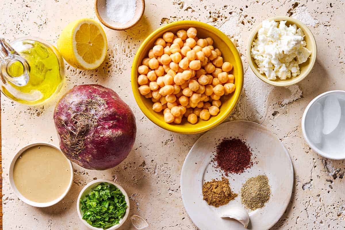 Ingredients for beet hummus including chickpeas, a beet, tahini paste, garlic, lemon, salt, cumin, coriander, sumac, olive oil, feta cheese, parsley and ice.