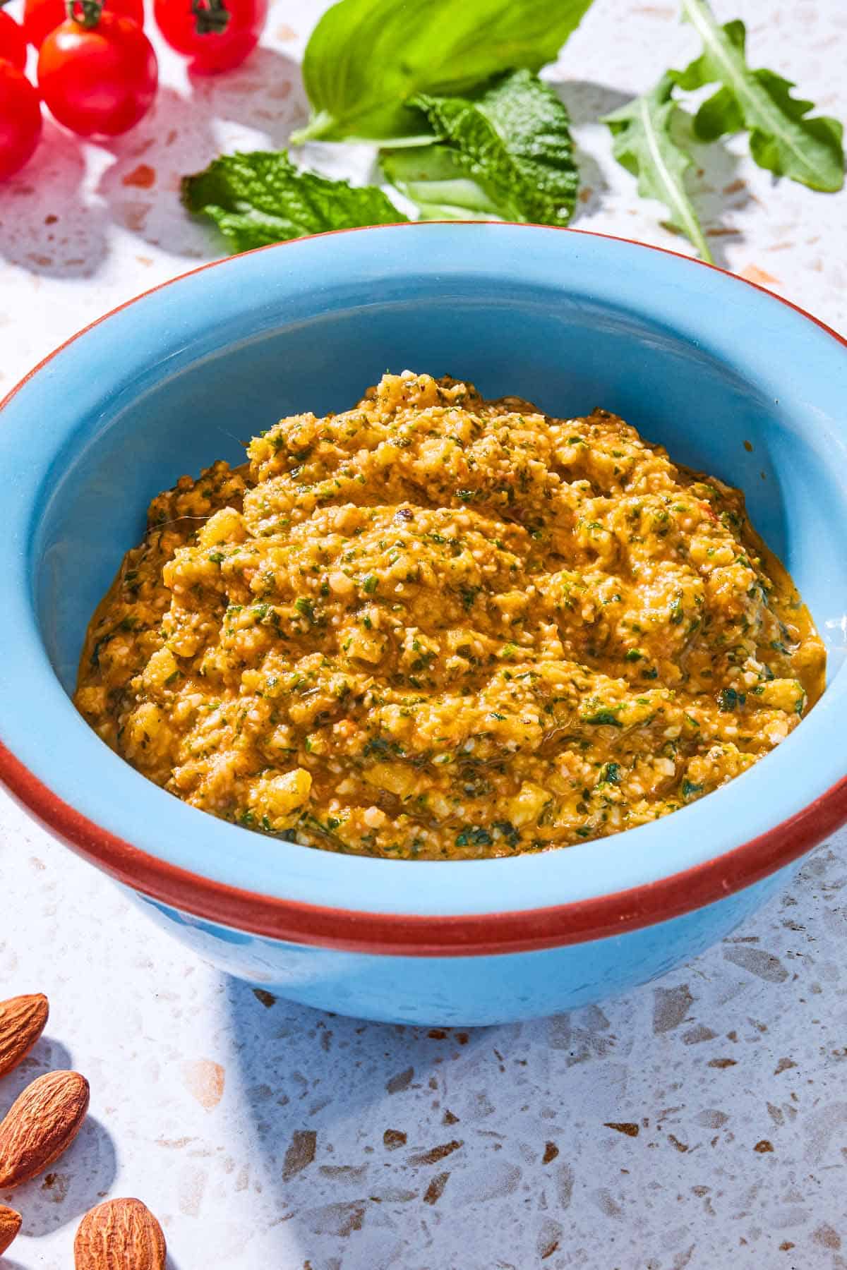 A close up of pesto alla trapanese in a bowl surrounded by cherry tomatoes, mint, arugula and almonds.