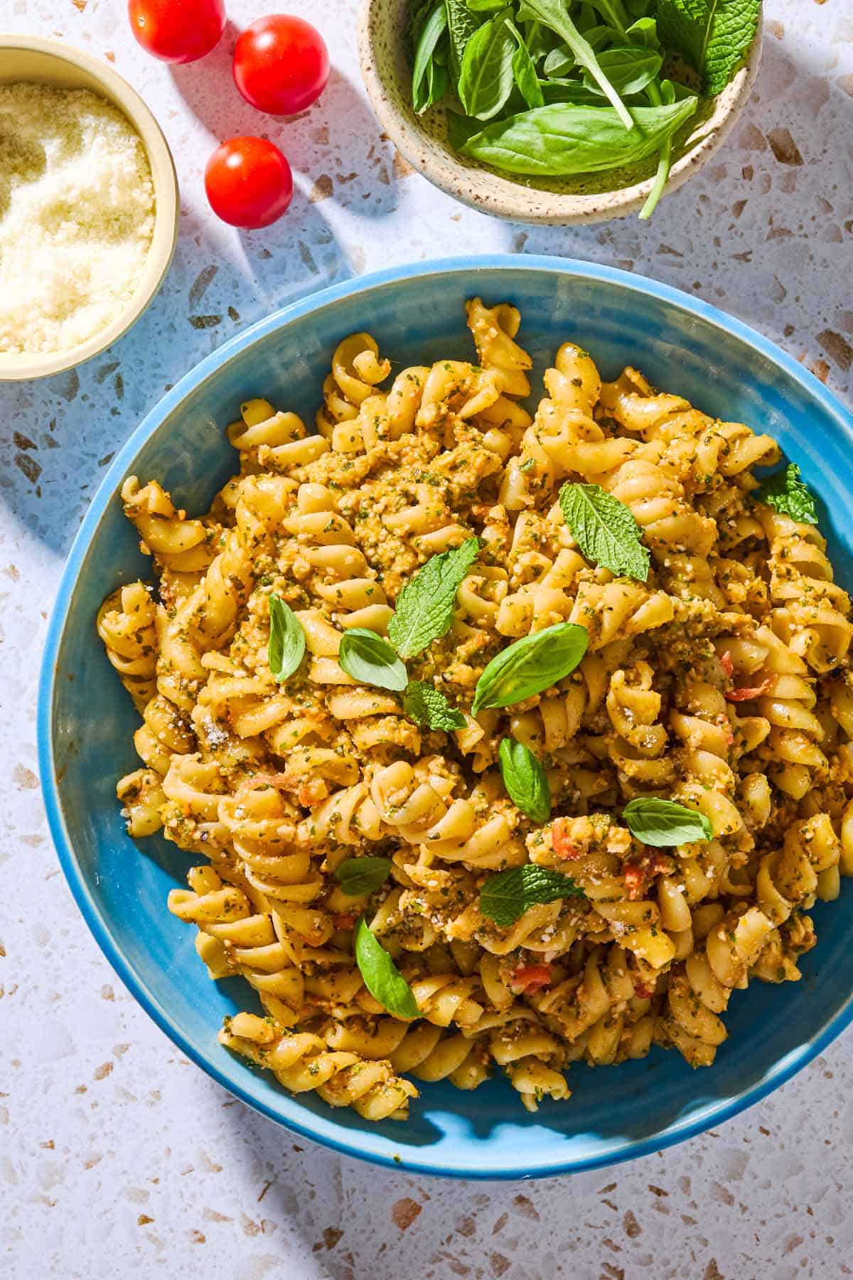 An overhead photo of pesto alla trapanese pasta in a bowl surrounded by 3 cherry tomatoes and bowls of pecorino romano cheese, and basil and mint.