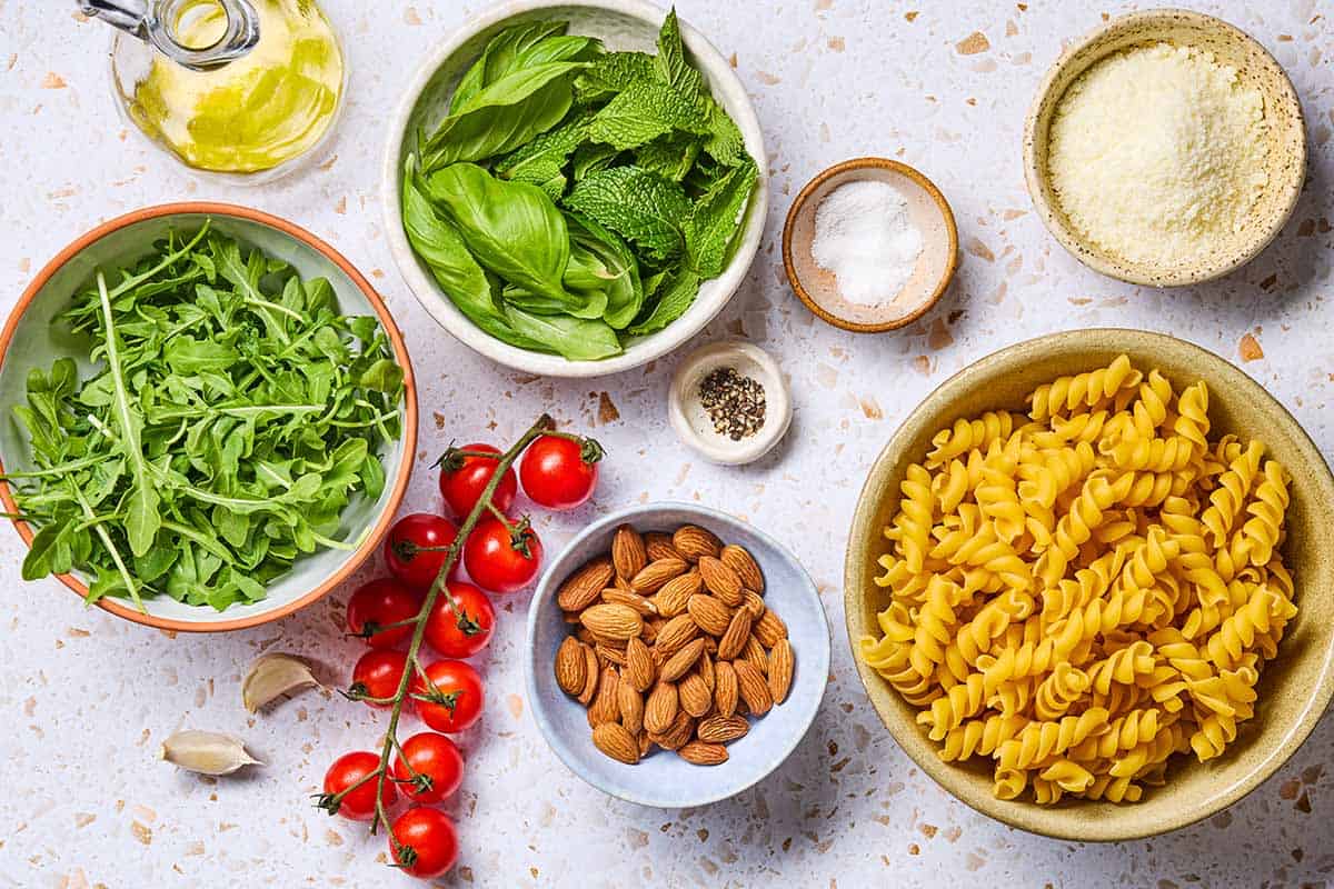 Ingredients for pesto alla trapanese including salt, cherry tomatoes, almonds, basil, mint, arugula, garlic, black pepper, olive oil, pecorino romano cheese, and pasta.