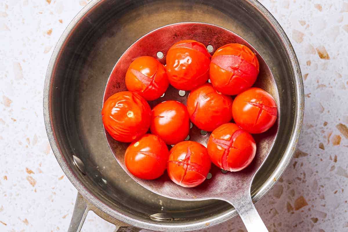 Several cherry tomatoes in a large slotted spoon being held over a saucepan of water, just after being blanched.