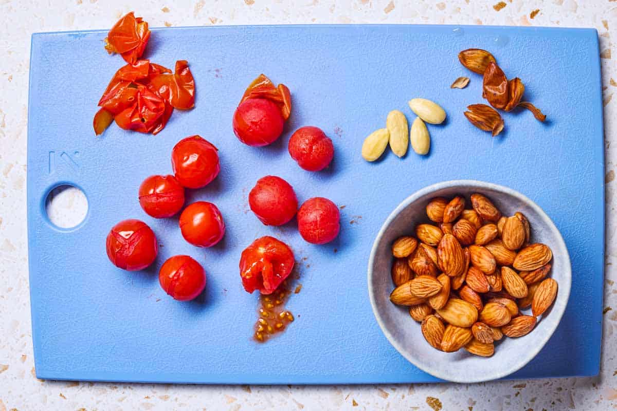 Several blanched tomatoes and almonds after being peeled on a cutting board. Next to these is a bowl of almonds with their peels.