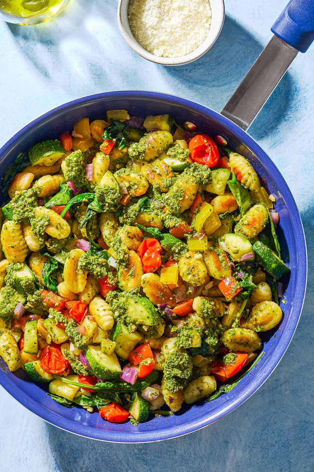 An overhead photo of crispy gnocchi with pesto in a skillet. Next to this is a bowl of grated parmesan cheese.