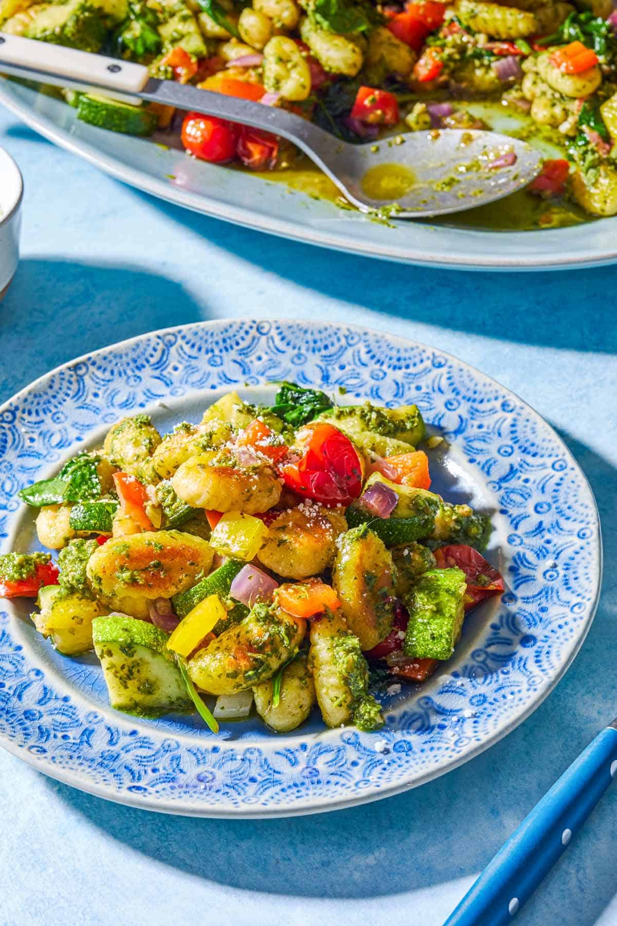 A close up of a serving of crispy gnocchi with pesto on a plate in front of the rest of the gnocchi on a serving platter with a spoon.