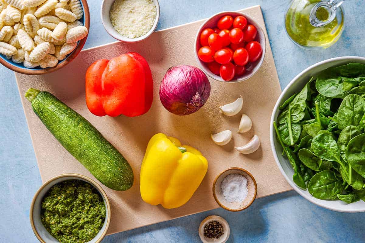 Ingredients for crispy gnocchi with pesto including gnocchi, olive oil, red onion, garlic, zucchini, red and yellow bell peppers, grape tomatoes, salt, pepper, baby spinach, basil pesto, and parmesan cheese.