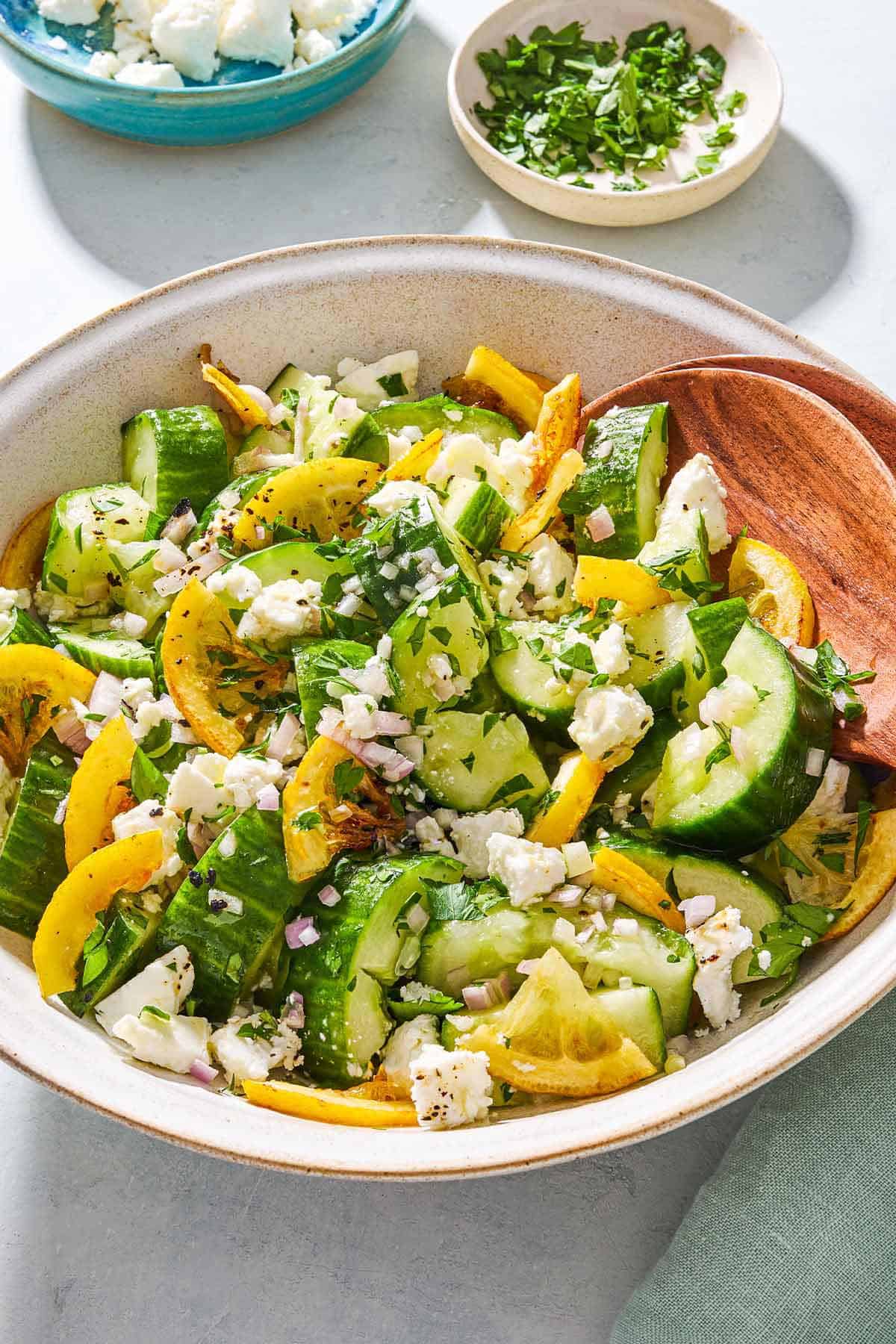 Smashed cucumber salad in a serving bowl with wooden serving utensils next to bowls of feta and parsley.