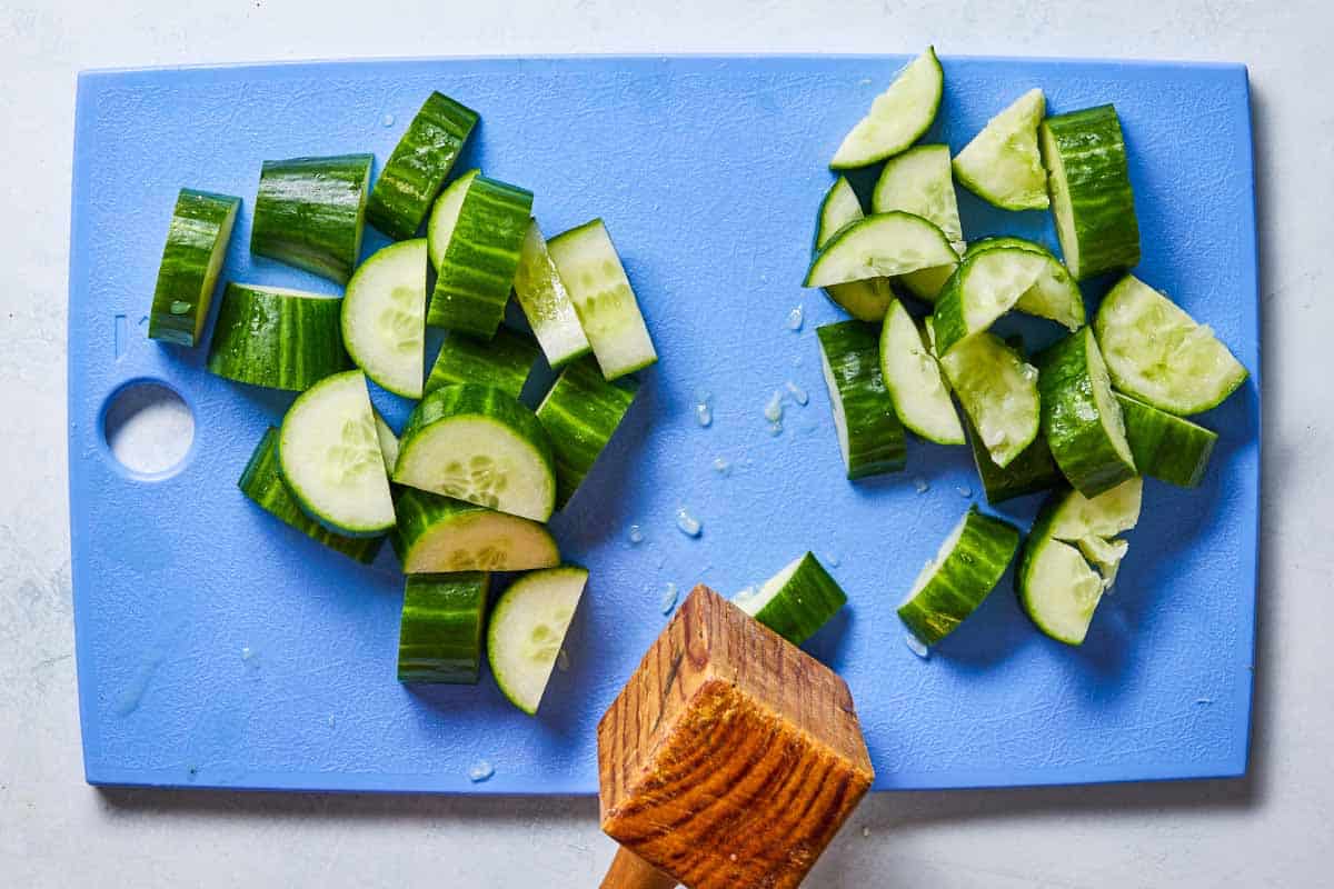 An overhead photo of cucumber slices on a cutting board. Half of them have been smashed with a wooden mallet.