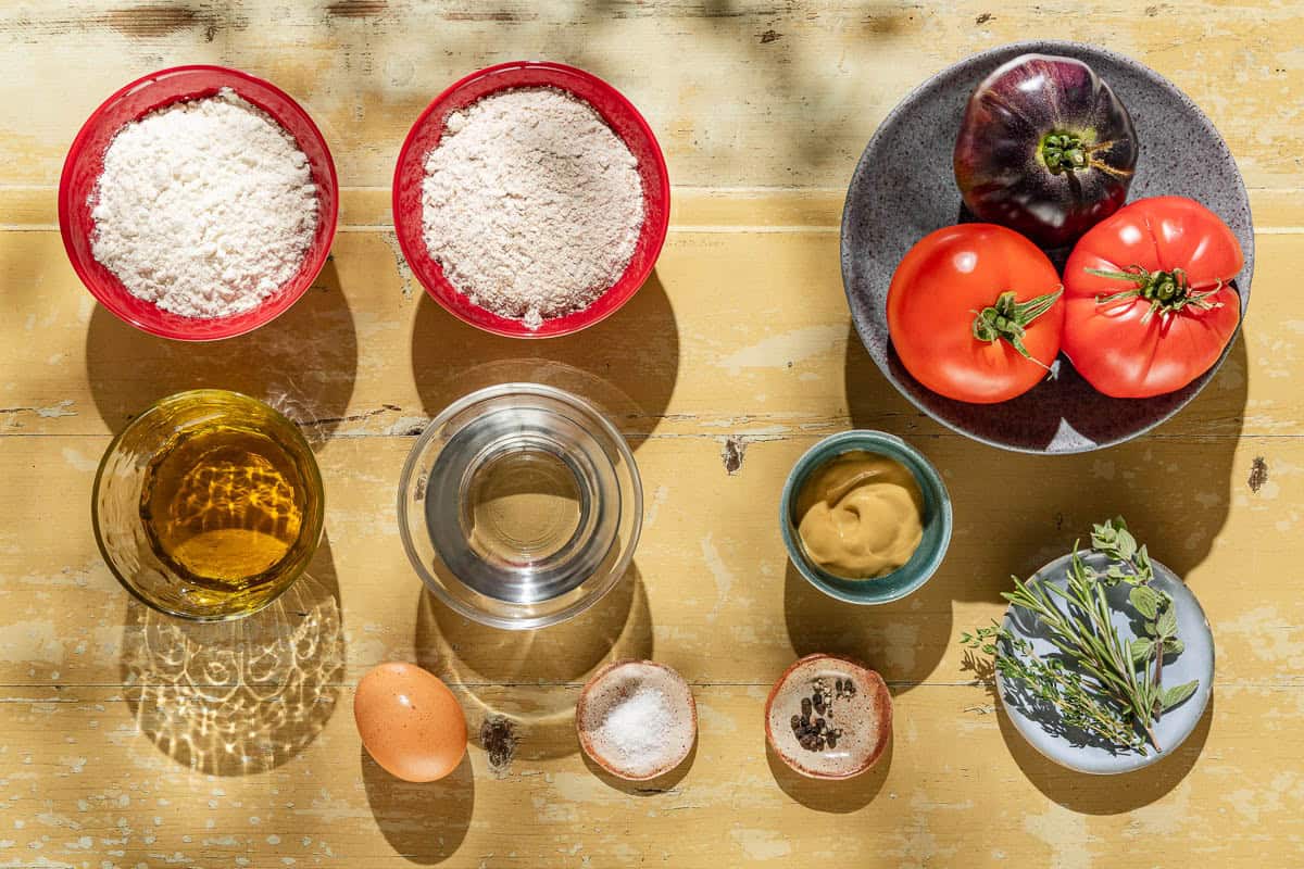 Ingredients for tomato tart including tomatoes all purpose flour, whole wheat flour, salt, olive oil, water, and egg, dijon mustard, black pepper, thyme, rosemary and marjoram.