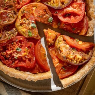An overhead photo of a tomato tart on a plate on a wooden cutting board with one slice cut from it. Next to this is a kitchen towel and a serving utensil.