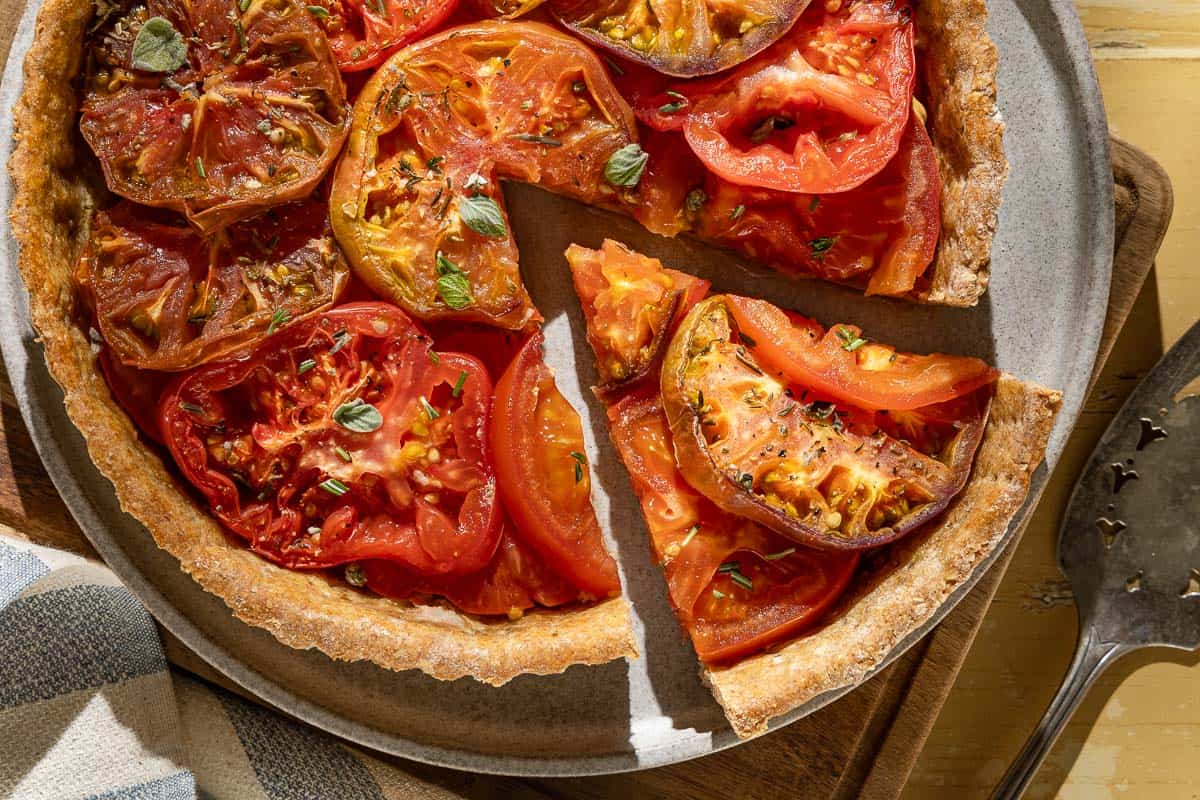 An overhead photo of a tomato tart on a plate on a wooden cutting board with one slice cut from it. Next to this is a kitchen towel and a serving utensil.