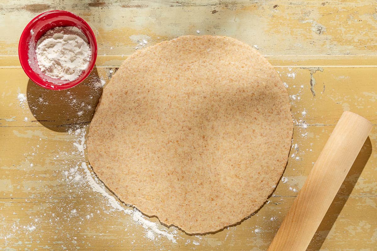 An overhead photo of the dough for the tomato tart rolled out on a table. Next to this is a small bowl of flour and a rolling pin.