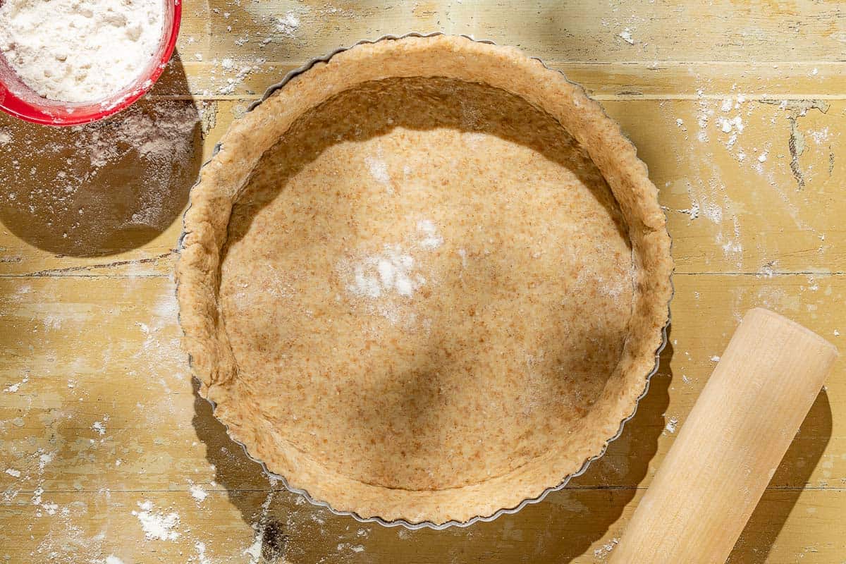 An overhead photo of the unbaked crust for the tomato tart. Next to this is a rolling pin and a bowl of flour.