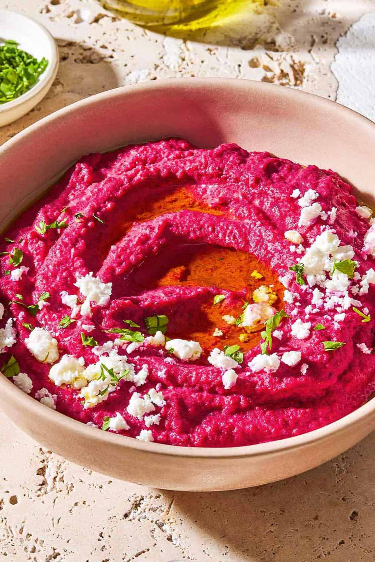 A close up photo of beet hummus in a bowl topped with olive oil, feta cheese and parsley.