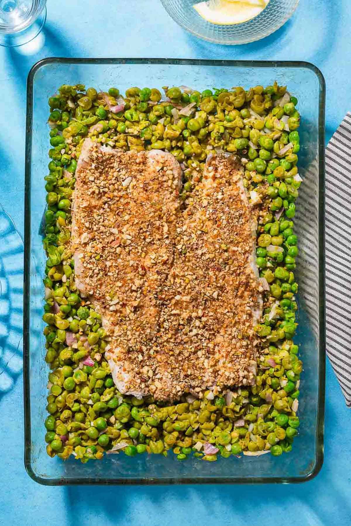 An overhead photo of baked white fish topped with dukkah on a bed of smashed peas in a baking dish.