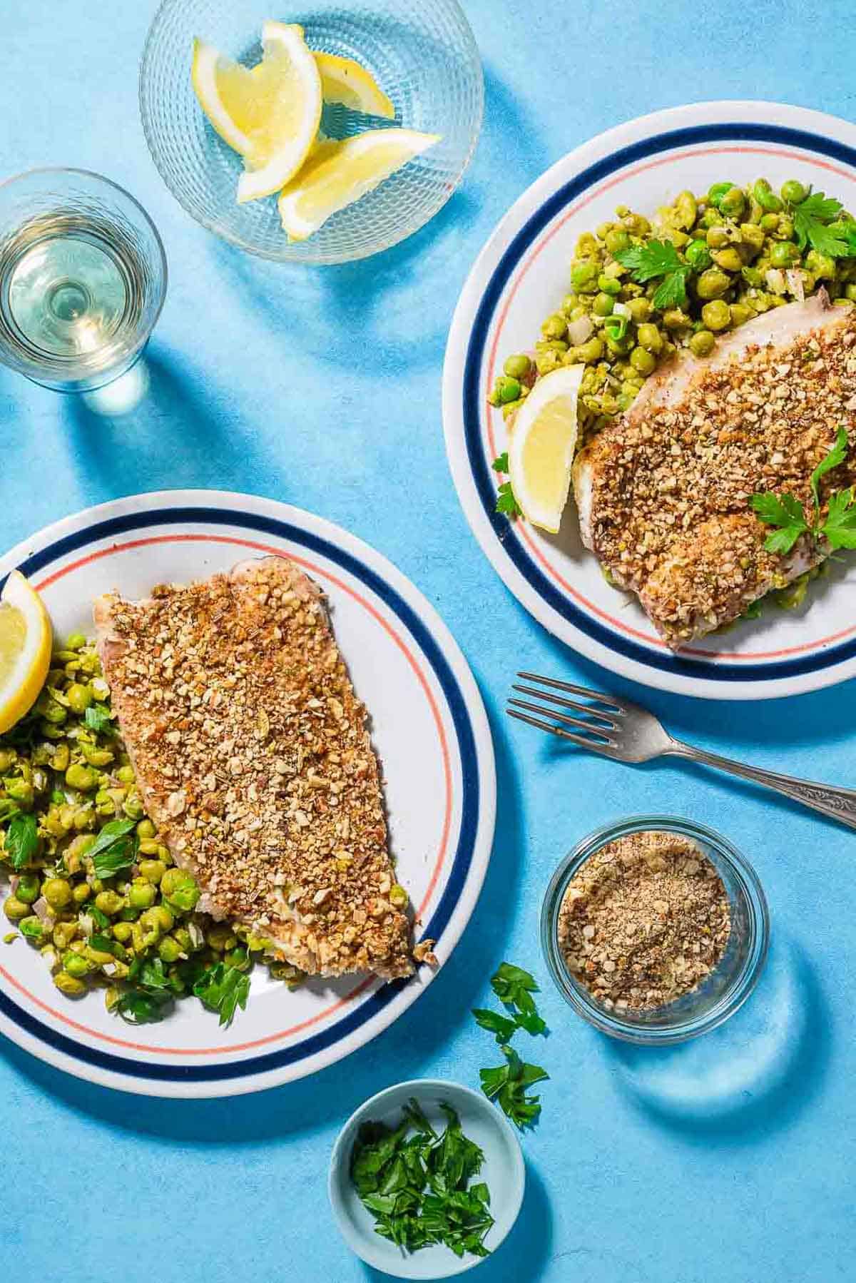 An overhead photo of 2 servings of baked white fish topped with dukkah on beds of smashed peas on plates with lemon wedges. Next to these are 2 forks, bowls of chopped parsley, dukkah and lemon wedges and a glass of wine.