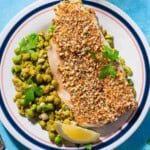 A close up of a serving of baked white fish toppedwith dukkah on a bed of smashed peas on a plate with a lemon wedge.