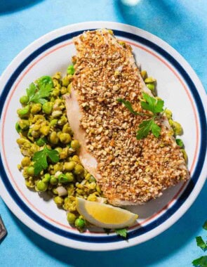 A close up of a serving of baked white fish toppedwith dukkah on a bed of smashed peas on a plate with a lemon wedge.