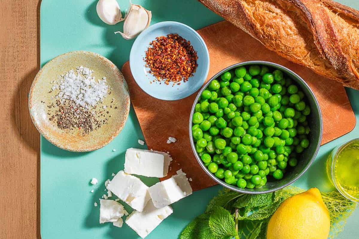 Ingredients for smashed peas on toast including shelled peas, olive oil, garlic, lemon, feta, mint, a baguette, aleppo pepper, salt, and black pepper.