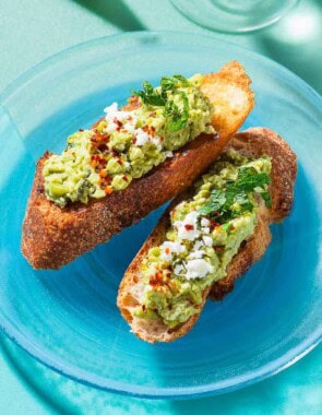 A close up photo of 2 pieces of smashed peas on toast garnished with aleppo pepper, feta and mint on a plate.