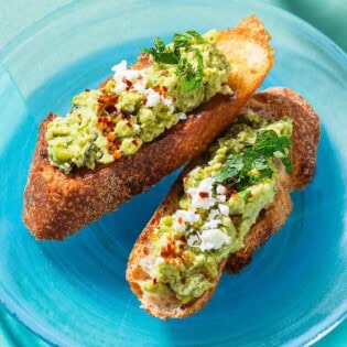 A close up photo of 2 pieces of smashed peas on toast garnished with aleppo pepper, feta and mint on a plate.